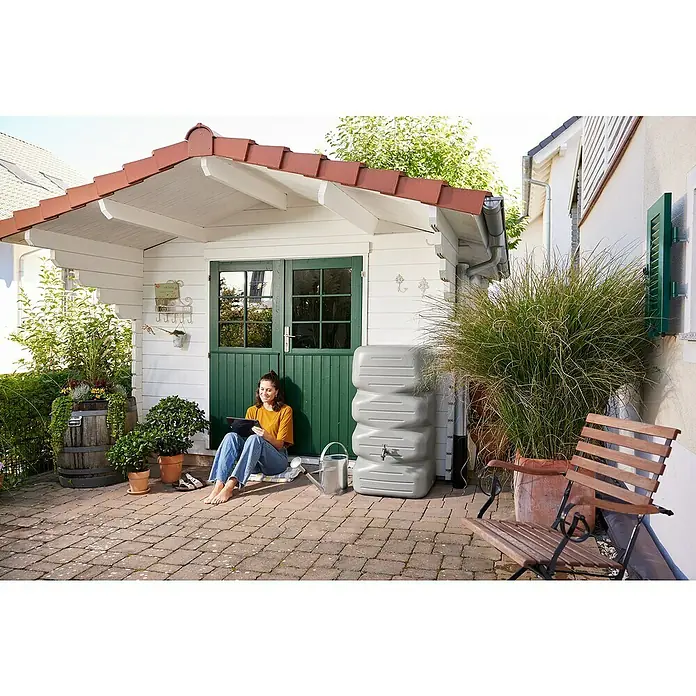 Garantia Basic Line Regentonne Cubus - 340 l, Betongrau - Eine Frau sitzt vor einem Gartenhaus mit einer grauen Wassersäule daneben, perfekt zur Gartenbewässerung und Wasserspeicherung.