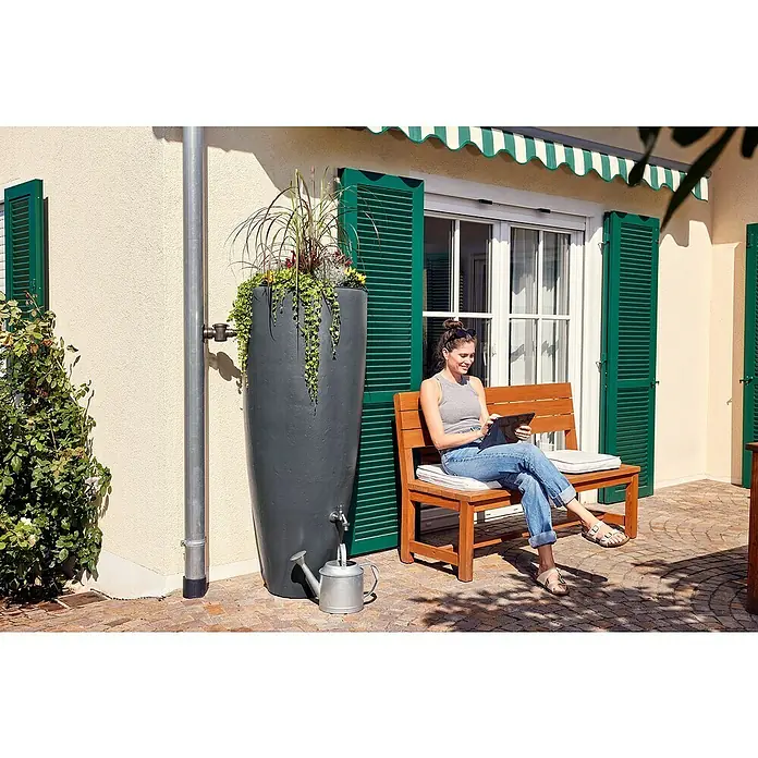 Garantia Classic Line Regenspeicher 2 in 1 - Graphite grey - Eine Frau sitzt auf einer Bank neben einer stilvollen Regenwassertonne, die mit Pflanzen geschmückt ist.