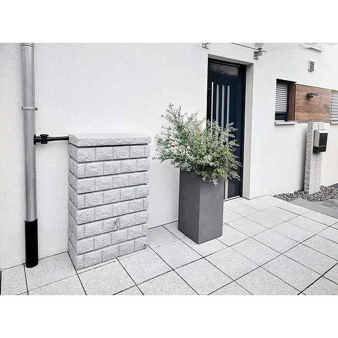 Garantia Exklusive Line Regenspeicher Rocky Junior - 300 l, Light Granit - Eine gemauerte Regentonne mit einem grauen Pflanzgefäß auf einer hellen, gefliesten Terrasse vor einem weißen Gebäude.
