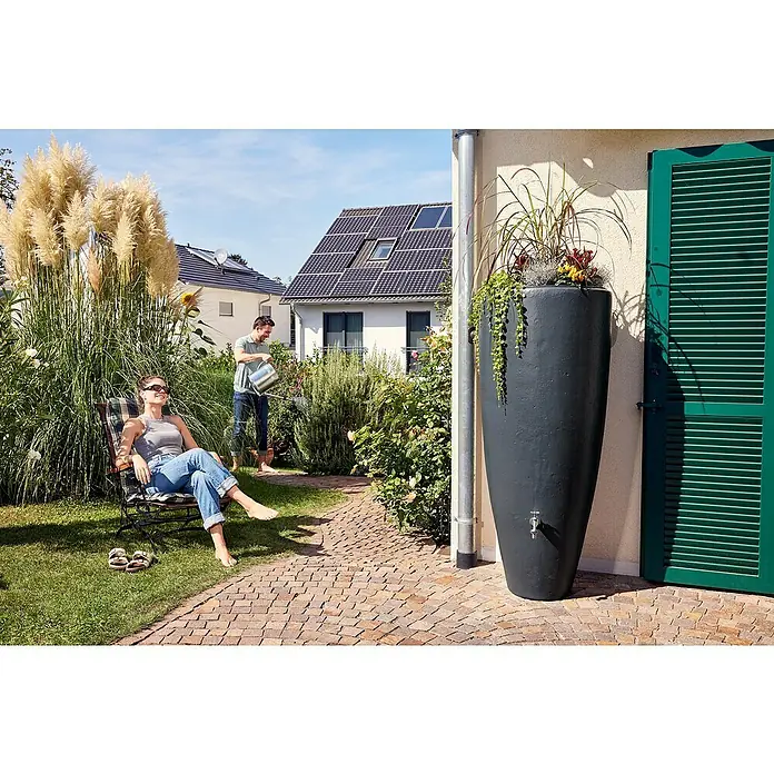 Garantia Classic Line Regenspeicher 2 in 1 - Graphite grey - Eine schlanke, graue Regentonnen-Säule mit Bepflanzung steht vor einer Hauswand. Im Hintergrund entspannt eine Frau, während ein Mann gießt.