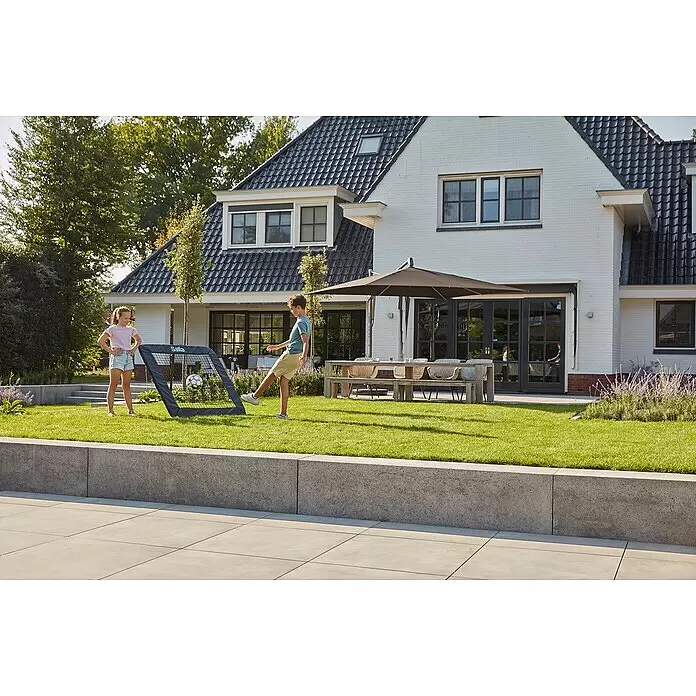 Salta Rebounder Motion - 124 x 124 cm, Schwarz - Dunkelgraues tragbares Salla Fußballtor, mit dem zwei Kinder in einem sonnigen Garten spielen. Ein Junge tritt den Ball ins Tor.