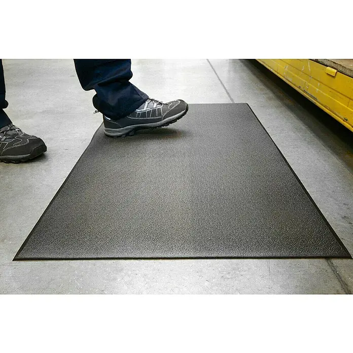 Coba Anti-Ermüdungsmatte Orthomat®-Standard - Breite: 90 cm, Schwarz - Eine Anti-Ermüdungsmatte in Schwarz, auf der ein Fuß steht, um den Komfort und die Sicherheit am Arbeitsplatz zu demonstrieren.