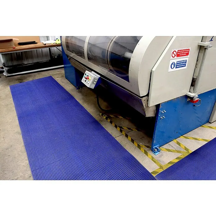 Coba Antirutschmatte DeckStepBlau, Breite: 100 cm, PVC - Eine blaue Bodenmatte mit Wabenmuster liegt vor einer großen Industriemaschine mit Warnhinweisen. Die Matte bietet Trittsicherheit und Komfort.