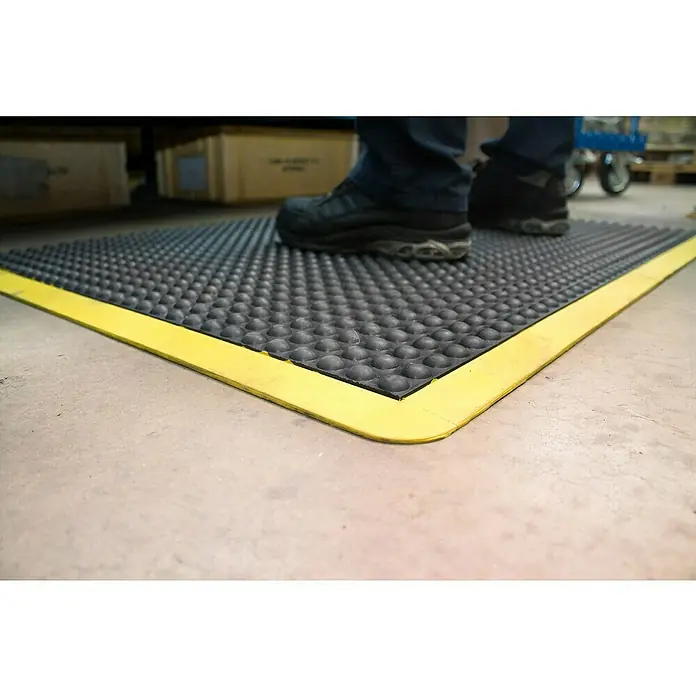 Coba Anti-Ermüdungsmatte Bubblemat Safety - 120 x 90 cm, Passend für: Coba Bubblemat Safety 28638689 / 28638670 - Schwarze Anti-Ermüdungsmatte mit gelbem Rand und Noppenoberfläche, auf der eine Person mit Arbeitsschuhen steht, für Komfort und Sicherheit.