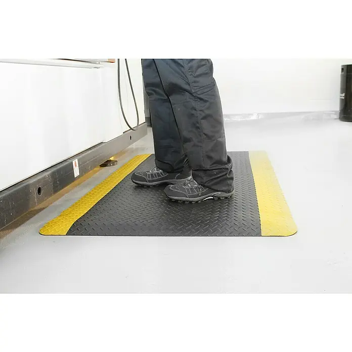 Coba Anti-Ermüdungsmatte Deckplate ProfiSafe Meterware - Breite: 90 cm, Rutschfest - Eine gelb-schwarze Antirutschmatte mit Riffelblechmuster, auf der eine Person mit Sicherheitsschuhen steht. Schützt vor Ausrutschen und Ermüdung.