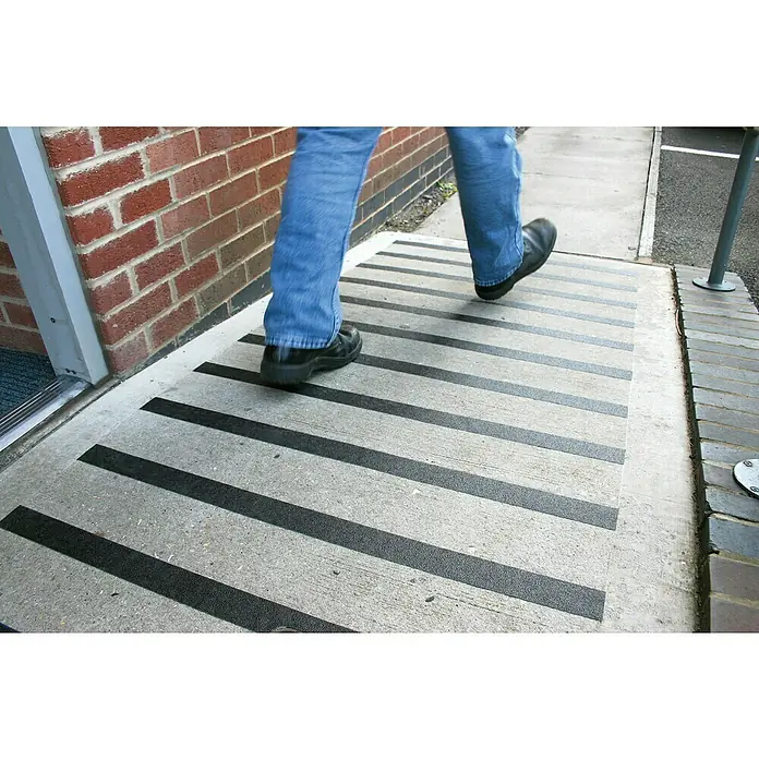 Coba Antirutschband Gripfoot Conformable - 183 x 2,5 cm, Schwarz - Nahaufnahme einer Person, die auf einer Betonrampe mit rutschfesten schwarzen Streifen geht, die zusätzliche Sicherheit bieten.