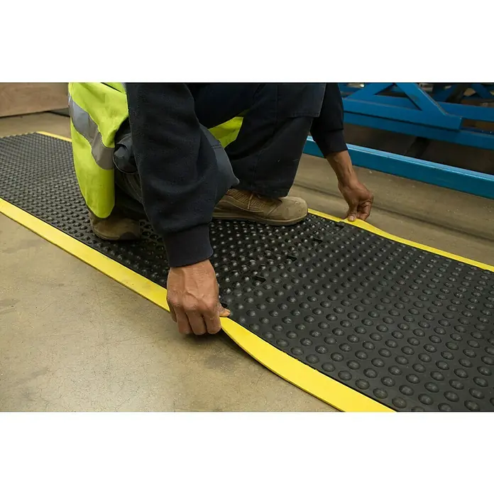 Coba Anti-Ermüdungsmatte Bubblemat Connect Ecke - 50 x 50 cm, Passend für: Coba Connect Safety 28638661 / 28638616 - Eine rutschfeste, schwarze Industriematte mit gelben Rändern wird auf einem Fabrikboden verlegt, um die Sicherheit zu erhöhen und Arbeitskomfort zu bieten.