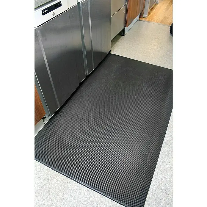 Coba Anti-Ermüdungsmatte Orthomat®-Ultimate90 x 60 cm, Schwarz - Eine schwarze, rechteckige Anti-Ermüdungsmatte liegt auf einem hellen Boden neben einer Edelstahl-Küchenzeile.