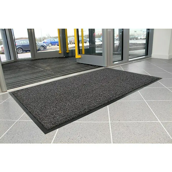 Coba Schmutzfangmatte CosmoSchwarz, 90 x 60 cm - Ein grauer Fußabtreter mit schwarzem Rand liegt vor einem Eingang mit Drehkreuz, der Schmutz und Nässe zuverlässig abfängt.