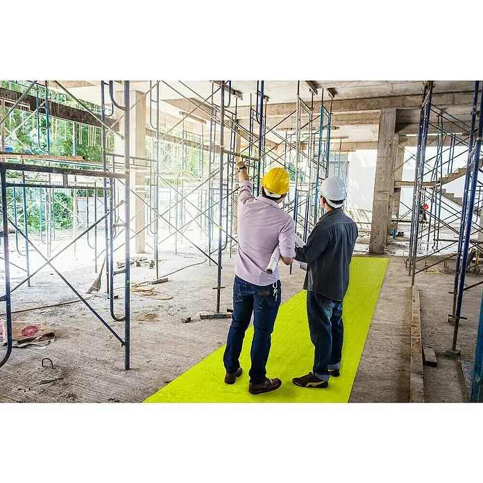 Coba Universalmatte SitePath - L x B: 10 x 1 m, Gelb, Einsatzbereich: Baustelle - Zwei Bauarbeiter mit Schutzhelmen stehen auf einer gelben Matte in einem Gebäude im Bau. Sie halten Pläne und diskutieren.