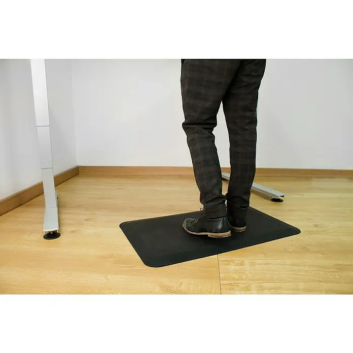 Coba Anti-Ermüdungsmatte Orthomat®-Office - 80 x 50 cm, Schwarz - Eine schwarze Stehmatte unter einem Schreibtisch mit einer Person, die darauf steht. Die Matte sorgt für Komfort und unterstützt die Ergonomie am Arbeitsplatz.