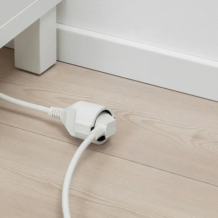 Chacon Alargador - 5 m, Blanco, H05VV-F - Primer plano de una regleta blanca con un enchufe insertado, sobre un suelo de madera clara junto a un rodapié blanco.