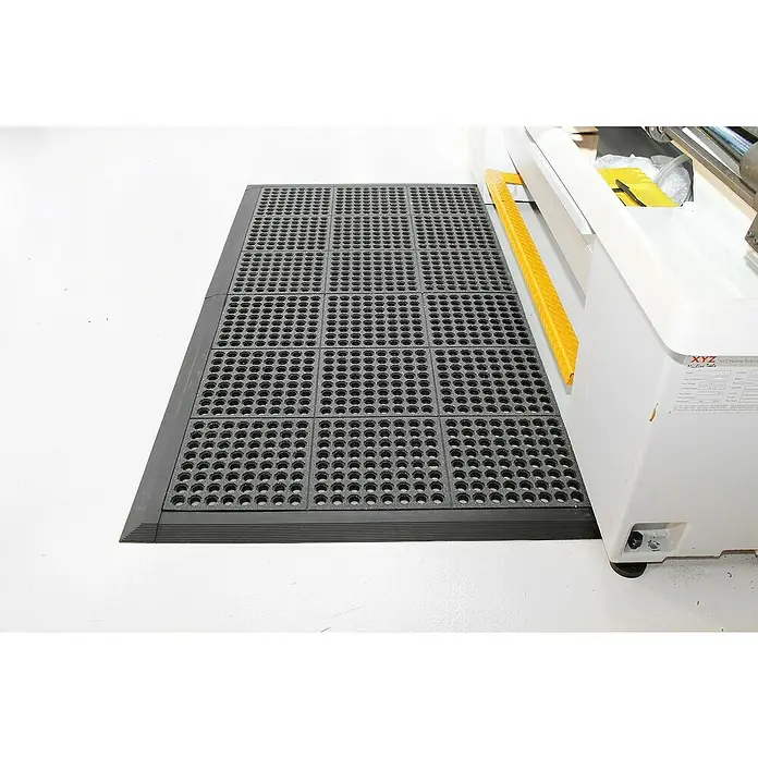 Coba Anti-Ermüdungsmatte Fatigue-Step Grit Top - 90 x 90 cm, Gelochte Oberfläche - Bild einer grauen, modularen Arbeitsplatzmatte mit Löchern und abgerundeten Kanten vor einer Werkbank.