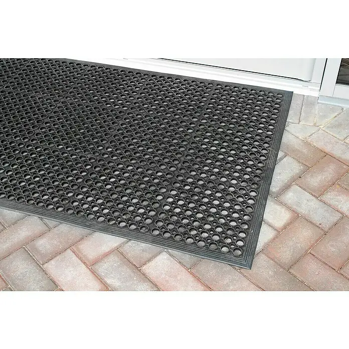 Coba Ringmatte Rampmat - 150 x 90 cm, Schwarz - Eine schwarze Gummimatte mit Löchern liegt vor einer Tür auf einem Pflasterweg. Die Matte ist robust und wetterfest.