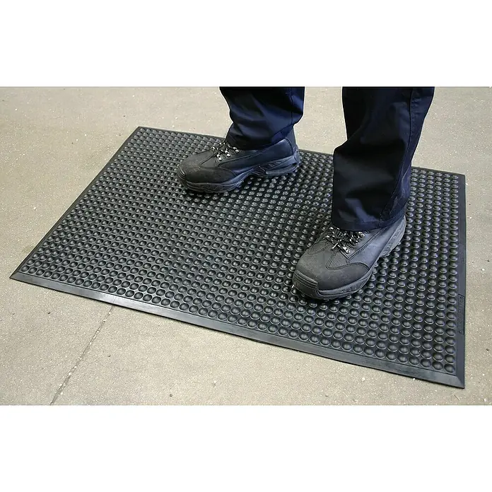 Coba Anti-Ermüdungsmatte Elite® Bubble90 x 120 cm - Eine schwarze Anti-Ermüdungsmatte mit Noppenstruktur, auf der eine Person mit schwarzen Schuhen steht, für Komfort und Sicherheit.