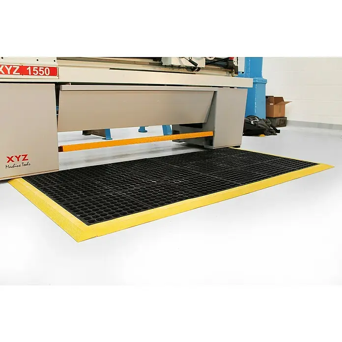 Coba Anti-Ermüdungsmatte Solid Fatigue-Step - Gelochte Oberfläche - Schwarze, wabenförmige Antirutschmatte mit gelbem Rand vor einer Werkzeugmaschine. Bietet sicheren Stand und reduziert Ausrutschgefahr.