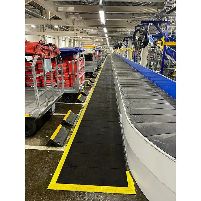 Coba Anti-Ermüdungsmatte Fatigue-Step90 x 90 cm, Einsatzbereich: Industrie - Eine Gepäckförderanlage mit Rollwagen voller roter Koffer in einem hellen Flughafenterminal. Gut sichtbar die Förderbandanlage.