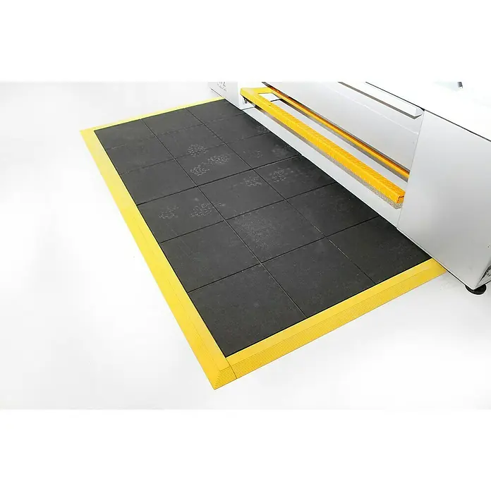 Coba Anti-Ermüdungsmatte Solid Fatigue-Step - Geschlossene Oberfläche - Schwarze Gummimatte mit gelbem Rand vor einer Maschine. Die Bodenmatte bietet Schutz und Sicherheit am Arbeitsplatz.