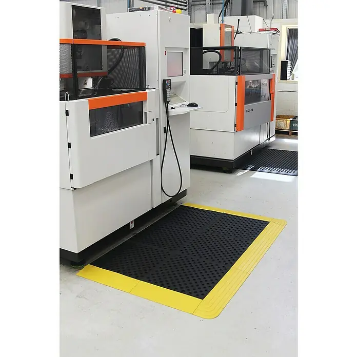Coba Eckelement Fatigue Lock Premium - Gelb, Passend für: Fatigue Lock 28977434 / 28977160 / 28977188 - Ein schwarze Antivermüdungsmatte mit gelbem Rand liegt vor Industriemaschinen und sorgt für Komfort und Sicherheit am Arbeitsplatz.