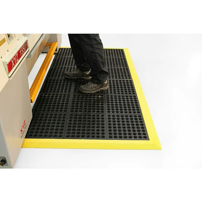 Coba Anti-Ermüdungsmatte Solid Fatigue-StepGelochte Oberfläche - Schwarze, modulare Anti-Ermüdungsmatte mit gelbem Rand, die auf dem Boden liegt und für Komfort und Sicherheit am Arbeitsplatz sorgt.