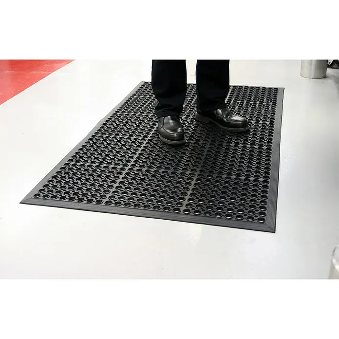 Coba Ringmatte Rampmat - 150 x 90 cm, Schwarz - Eine schwarze, perforierte Gummimatte liegt auf einem weißen Boden. Eine Person mit schwarzen Schuhen steht auf der Matte.