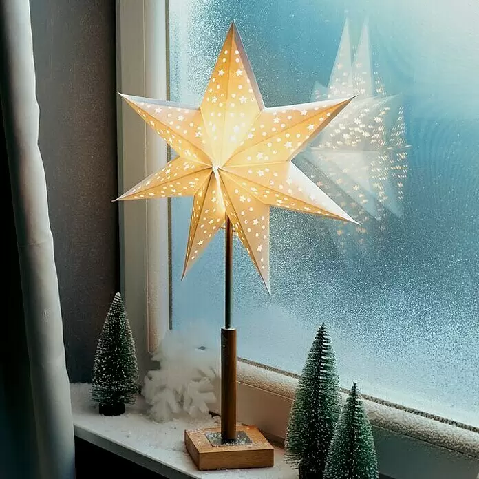 Markslöjd Standstern Solvalla - Weiß, Netzbetrieben - Beleuchtete sternförmige Papierlaterne mit Musterausschnitten auf einer verschneiten Fensterbank mit Miniatur-Bäumen.