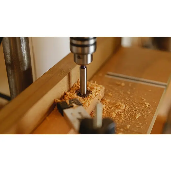 Bosch Taladro atornillador percutor de batería AdvancedImpact 18 - 18 V, 2 baterías, 1,5 Ah - Primer plano de un taladro de banco haciendo un agujero en un trozo de madera, creando virutas. La herramienta parece robusta y precisa.