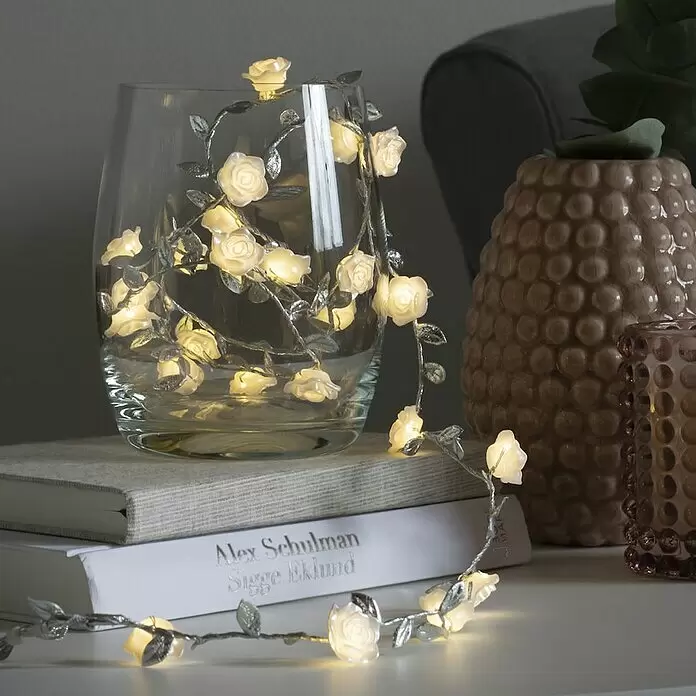 Konstsmide LED-Lichterkette - Innen, Lichtfarbe: Warmweiß, Anzahl Leuchtmittel: 25 Stk., Batteriebetrieben - Weiße LED-Rosenlichterkette mit silbernen Blättern, dekorativ in einer Glasvase und über Büchern drapiert.