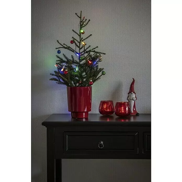 Konstsmide LED-Lichterkette Bunt - Lichtfarbe: Mehrfarbig, Anzahl Leuchtmittel: 35 Stk., Netzbetrieben, IP20 - Kleiner, dekorierter Weihnachtsbaum in rotem Topf mit festlichen roten Kerzenhaltern und einem Wichtel auf einem dunklen Tisch.