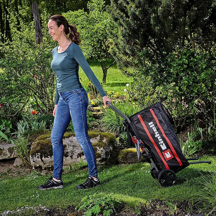 Einhell Transportwagen - L x B x H: 535 x 531 x 1.015 mm - Eine Frau zieht einen schwarzen Einhell-Gartenwagen mit roten Streifen durch einen grünen Garten.