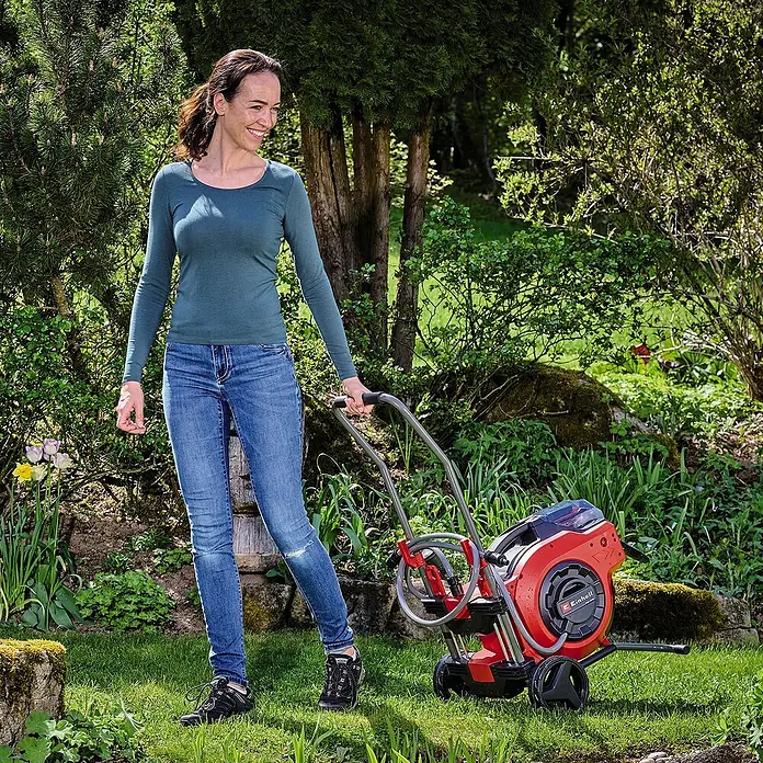 Einhell Transportwagen - L x B x H: 535 x 531 x 1.015 mm - Frau schiebt eine rote Einhell Schlauchbox mit Rädern durch den Garten. Praktisch für die Bewässerung.