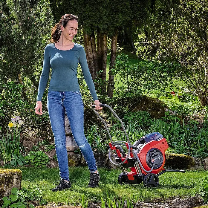 Einhell Power X-Change 18V Akku-Schlauchtrommel GE-HR 18/30 - 18 V, Ohne Akku - Frau zieht einen roten Einhell-Hochdruckreiniger auf Rädern über einen Rasen. Das Gerät ist kompakt und benutzerfreundlich gestaltet.