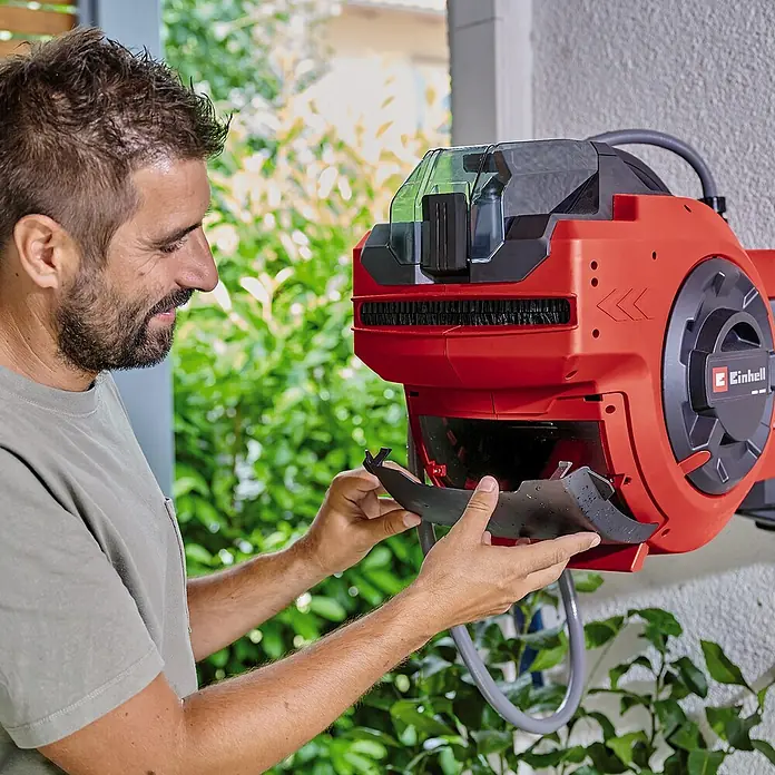 Einhell Power X-Change 18V Akku-Schlauchtrommel GE-HR 18/30 WH Li-Solo - 18 V, Ohne Akku, Schlauchkapazität: 30 m, 12,5 mm (½″) - Ein Mann inspiziert eine rote Einhell Schlauchtrommel an einer Wand. Das Gerät ist für die ordentliche Aufbewahrung von Gartenschläuchen konzipiert.