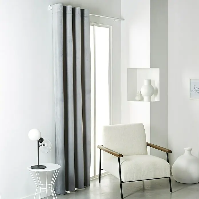 Verdunkelungsvorhang VelvetLight Grey, 135 x 245 cm, 100 % Polyester - Graue Vorhänge im Zimmer mit weißem Sessel, Tisch und Dekorationen. Moderner Look mit sanften Farben und elegantem Design für ein stilvolles Zuhause.