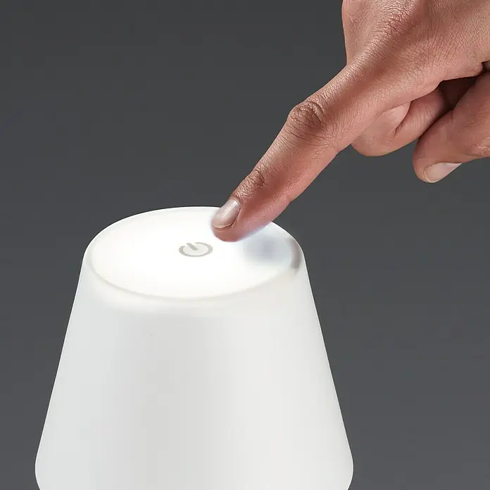FHL easy Akku-Tischleuchte TROPEA - 2,5 W, L x B x H: 12 x 12 x 38 cm, Sandweiß, Warmweiß - Nahaufnahme einer Person, die einen weißen Nachttischlampe mit einem Finger berührt, um sie einzuschalten.