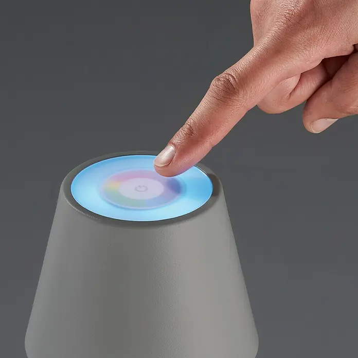 FHL easy Akku-Tischleuchte COSENZA - 2 W, L x B x H: 12 x 12 x 38 cm, Sand, RGB, 1 Stk. - Nahaufnahme eines grauen Nachttisch-Diffusors mit Farblicht und einem Finger, der ihn berührt.