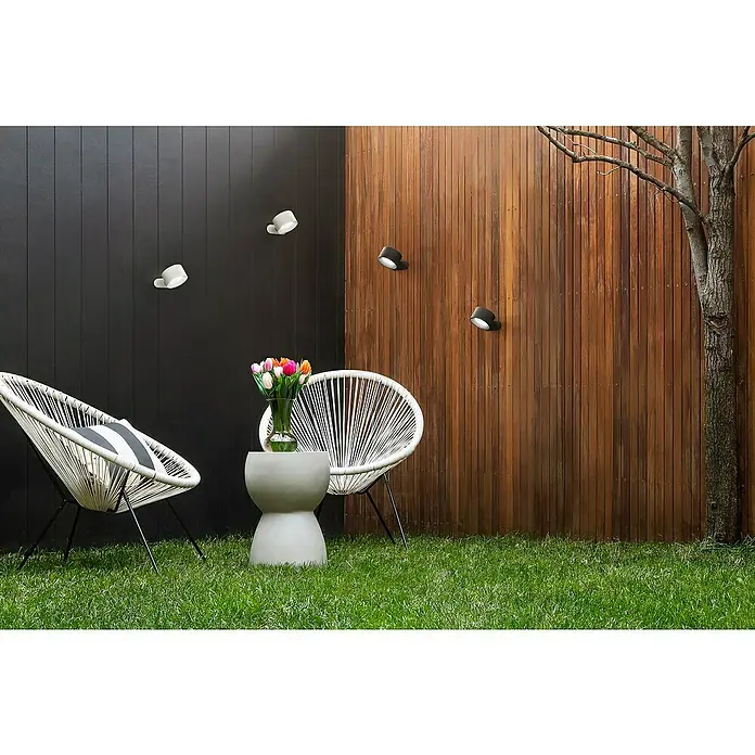 FHL easy Magnetic Akku-Wandleuchte9,5 x 8 x 4,5 cm, Weiß, Mehrfarbig, 2 W, IP20 - Gartenambiente mit weißen und schwarzen Wandleuchten an einer Holzvertäfelung, ergänzt durch moderne Korbsessel und einen Beistelltisch mit Blumen.