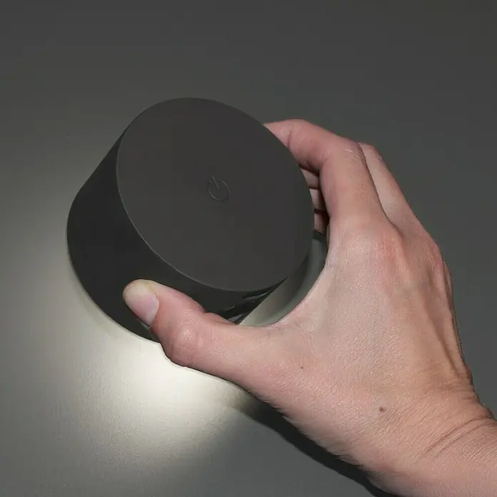 FHL easy Magnetic Akku-Wandleuchte - 9,5 x 8 x 4,5 cm, Schwarz, Mehrfarbig, 2 W, IP20 - Eine Hand hält einen grauen, zylindrischen Smart Speaker mit einem Power-Symbol auf der Oberseite vor einem neutralen Hintergrund.