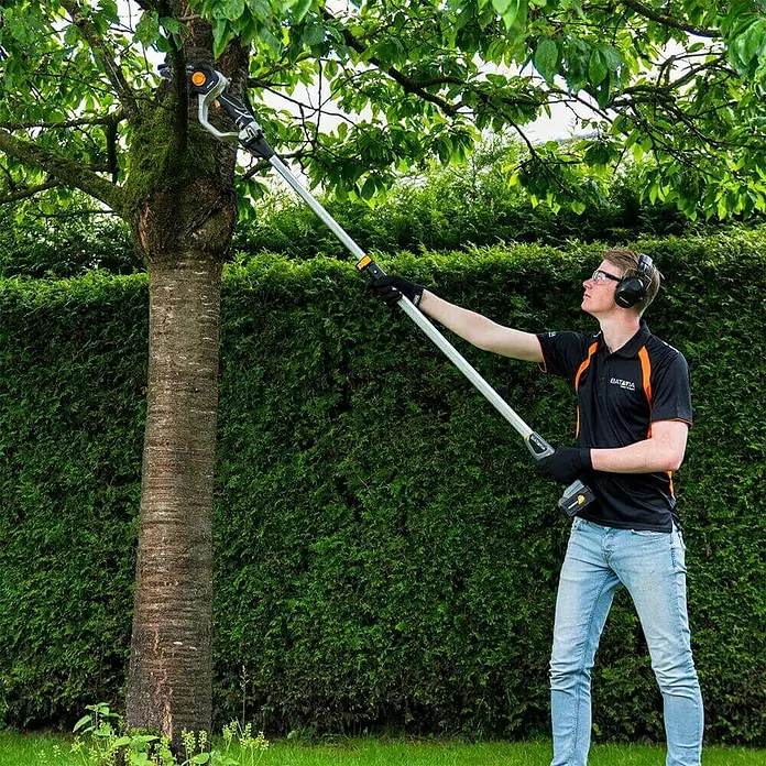Teleskopstiel - Passend für: Batavia Akku-Kettensäge Nexxsaw V3.1 & V2 - Ein Mann benutzt eine Akku-Astsäge, um Äste an einem Baum zu schneiden, mit einer hohen Hecke im Hintergrund.