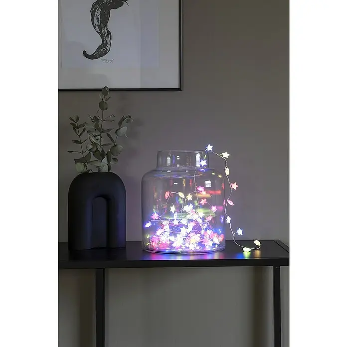 Konstsmide LED-Lichterkette - Innen, Lichtfarbe: Mehrfarbig, Anzahl Leuchtmittel: 90 Stk., Netzbetrieben - Eine sternförmige Lichterkette leuchtet in einem Glasgefäß auf einem schwarzen Regal, neben einer Vase mit Blättern und einem gerahmten Bild.