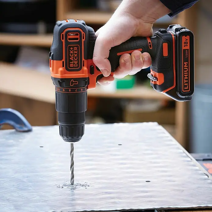 Black+Decker Taladro atornillador percutor de batería BDCHD18C2TA -  - Un taladro inalámbrico Black+Decker perforando una lámina de metal. El taladro, parte de un juego de herramientas, está sujeto por una mano.