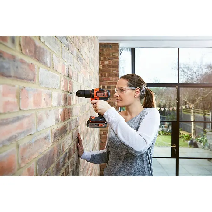 Black+Decker Taladro atornillador percutor de batería BDCHD18C2TA -  - Una mujer usa un taladro inalámbrico Black+Decker de 18 V en una pared de ladrillos. El taladro es naranja y negro y tiene una batería de 18 V.