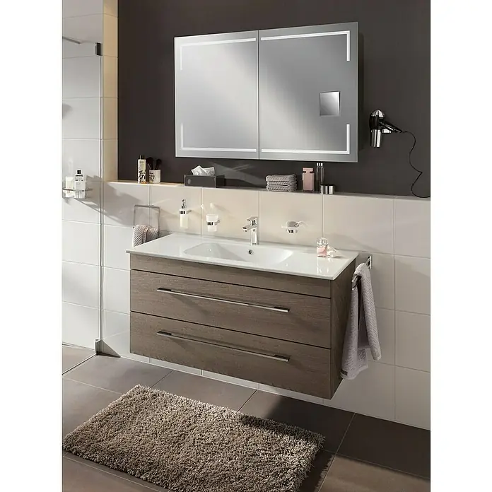 Lanzet K5 Waschtischunterschrank - 117,9 x 50,3 x 57,5 cm, Eiche Maron - Badezimmer-Interieur mit einem modernen Waschtischunterschrank in Holzoptik, einem beleuchteten Spiegelschrank und einem flauschigen Teppich.