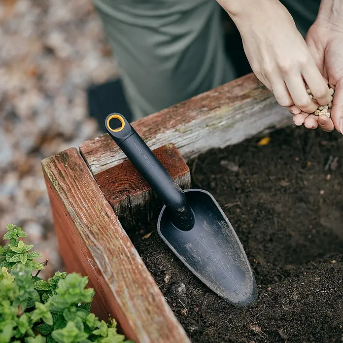 Fiskars Solid Blumenkelle - 28,2 cm - Nahaufnahme eines Fiskars-Pflanzspatens, der auf einem Hochbeet liegt, während jemand Samen in die Erde sät.