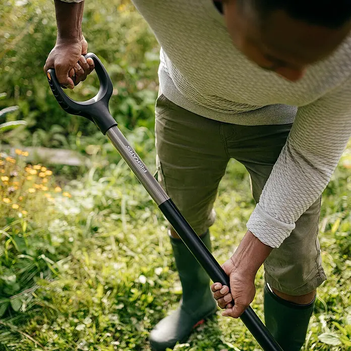 Fiskars Ergonomic Spatengabel - Arbeitsbreite: 20,5 cm, Anzahl Zinken: 4, Ergonomischer Stiel - Nahaufnahme einer Person mit einem Fiskars-Spaten im Garten, ideal für Gartenarbeiten und Landschaftsbau.