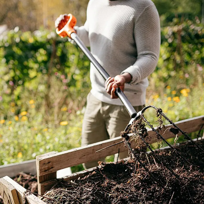 Fiskars Solid Dunggabel - Anzahl Zinken: 4, Langstiel - Eine Person benutzt eine Grabgabel mit orangem Griff, um Kompost in einem Garten umzugraben. Das Werkzeug erleichtert die Gartenarbeit.
