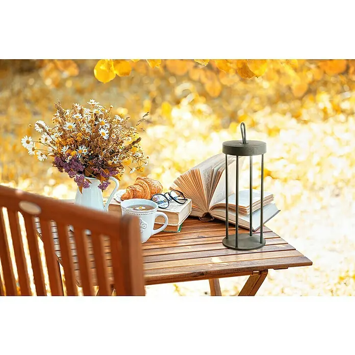 FHL easy Akku-Tischleuchte KANTE - 3 W, L x B x H: 13 x 13 x 36 cm, Schwarz, Warmweiß - Stillleben im Freien mit einer modernen Laterne auf einem Holztisch, umgeben von Blumen, Büchern und einem Croissant.