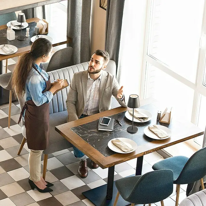 FHL easy Akku-Tischleuchte COSENZA 2.0 - 2 W, L x B x H: 11 x 11 x 34 cm, Sandschwarz, RGB - Eine Restaurant-Szene mit einem Tisch, auf dem eine elegante Tischlampe steht, während ein Kellner die Bestellung eines Kunden aufnimmt.