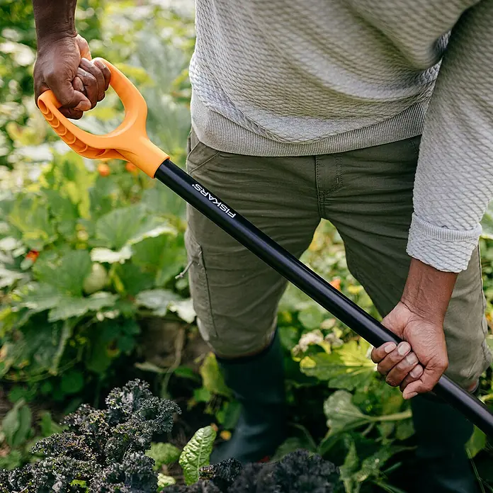 Fiskars Solid Dunggabel - Anzahl Zinken: 4, Langstiel - Nahaufnahme einer Person mit einem Fiskars-Gartengerät im Garten. Die orangefarbenen Akzente des Werkzeugs stechen hervor.