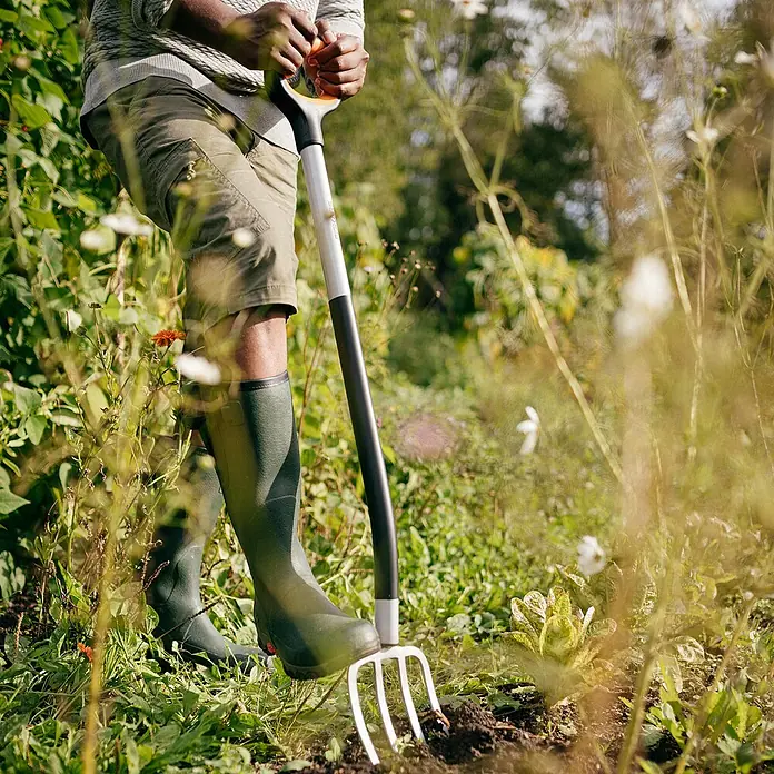 Fiskars Xact Spatengabel - Arbeitsbreite: 18,5 cm, Anzahl Zinken: 4, Langstiel - Auf dem Bild ist eine Gartenforke vor dem Hintergrund eines Gärtners in Stiefeln zu sehen, ideal für Gartenarbeiten und Bodenbearbeitung.